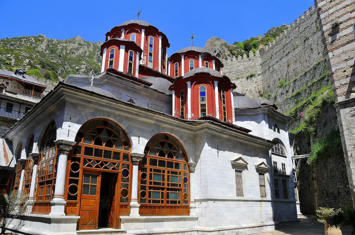 The white marble main church of Saint Paul Monastery on Mount Athos with distinct red domes, wooden framed windows, and a stone defensive wall in the background under a blue sky.
