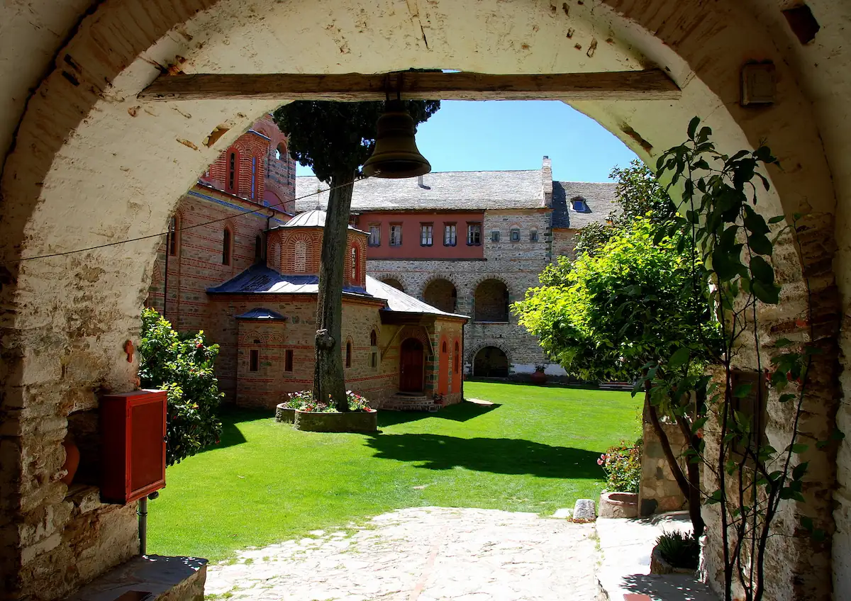 The inner courtyard of Filotheou Monastery on Mount Athos, featuring the central church, a green lawn, and a stone archway in the foreground.