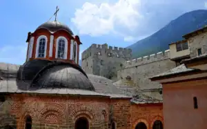 Courtyard of Great Lavra Monastery in Mount Athos, featuring a Byzantine church with a red and lead dome, and a medieval fortified stone tower against a mountain background.