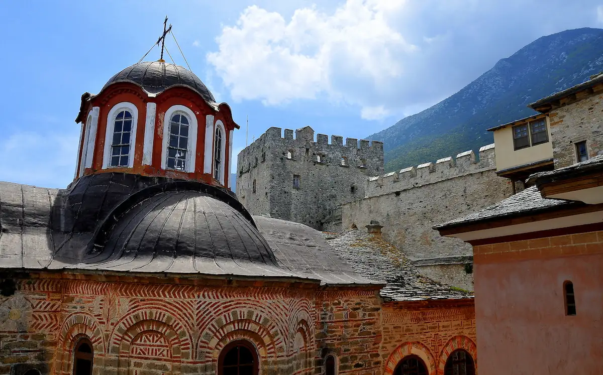 Courtyard of Great Lavra Monastery in Mount Athos, featuring a Byzantine church with a red and lead dome, and a medieval fortified stone tower against a mountain background.